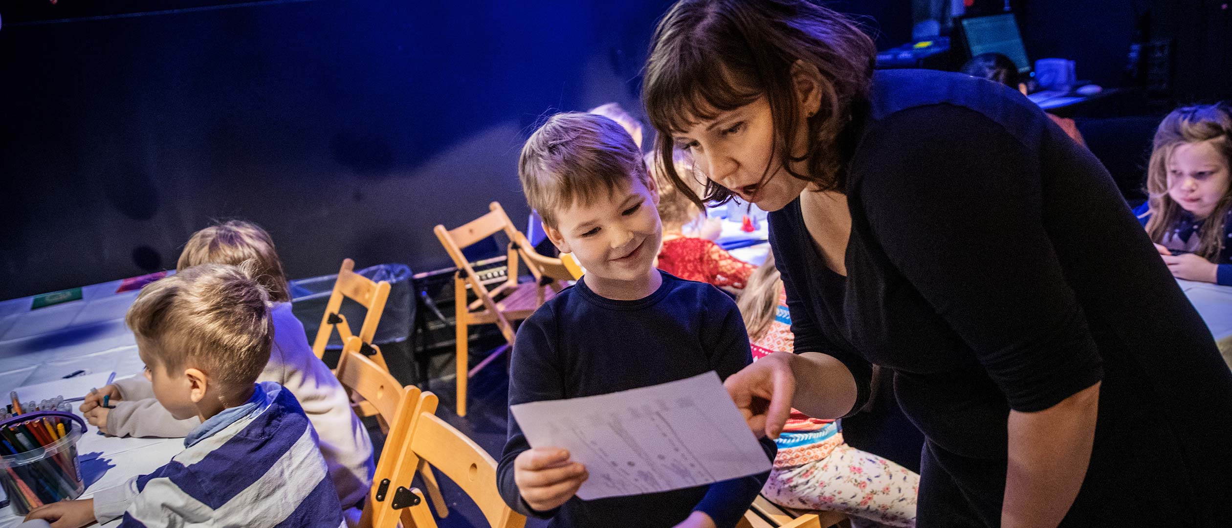 Barn sitter vid bord och ritar. Ett barn visar sin teckning åt en pedagog.