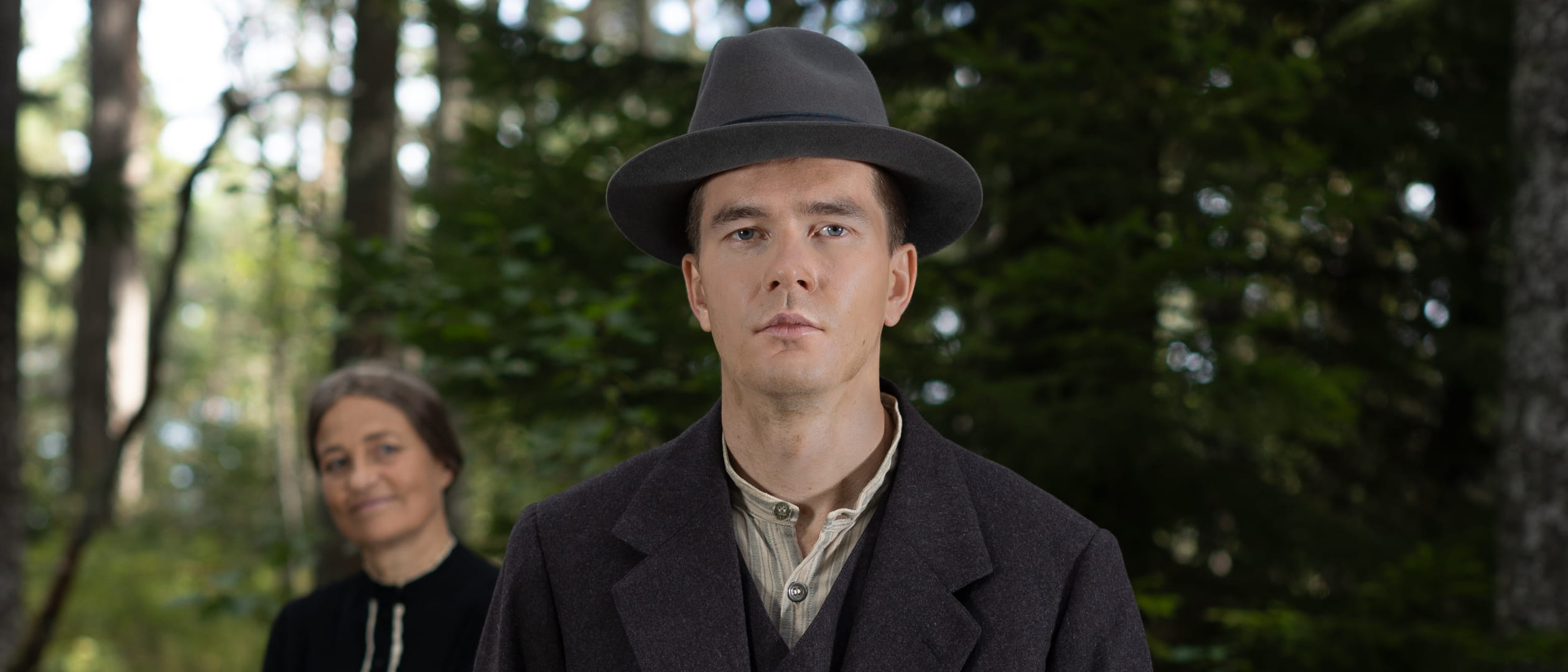Frans Lejon står i en skog och ser framåt med ett allvarligt ansiktsuttryck. En kvinna ser på honom med ett leende i bakgrunden.
