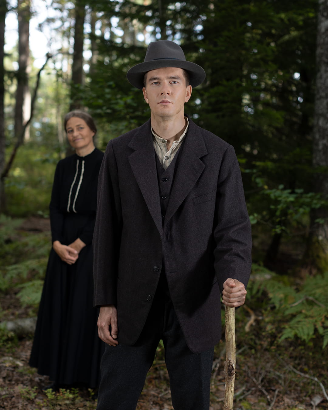 Frans Lejon står i en skog och ser framåt med ett allvarligt ansiktsuttryck. En kvinna ser på honom med ett leende i bakgrunden.